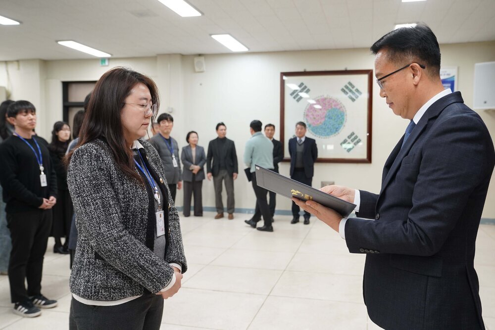 2026년 상반기 4급이하 정기인사로 정장을 입은 남성이 실내 행사장에서 한 여성에게 상장을 전달하며 내용을 읽고 있고, 여성은 회색 재킷을 입고 두 손을 모은 상태로 서 있으며 뒤쪽에는 참석자들이 줄지어 서서 행사를 지켜보고 있다.