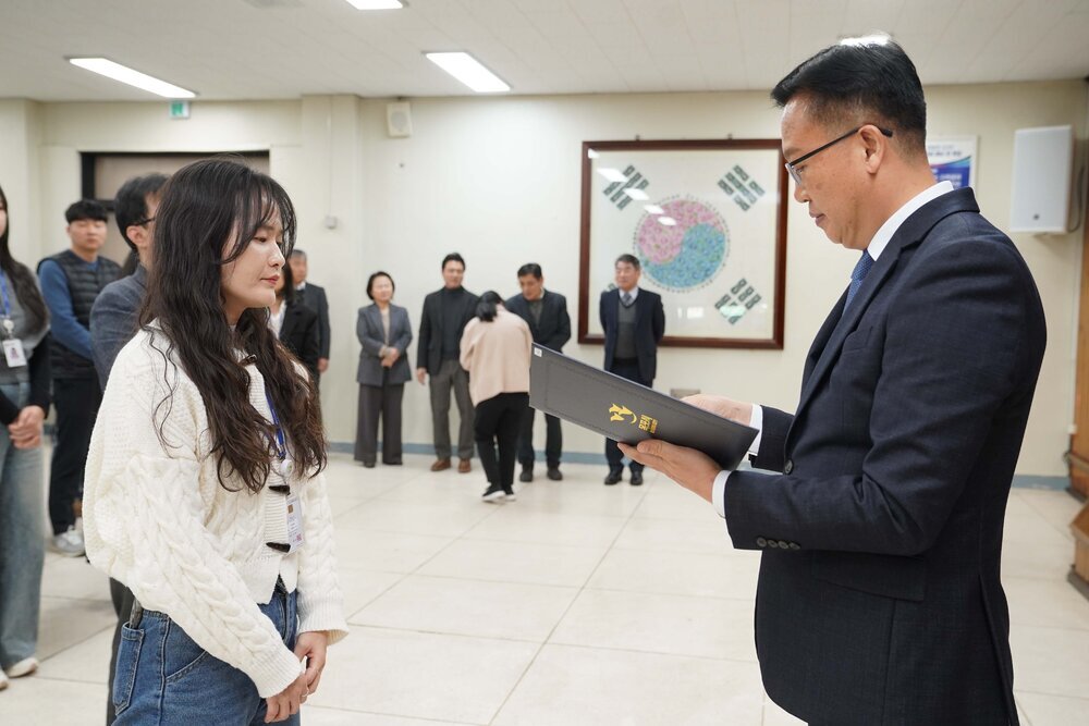 2026년 상반기 4급이하 정기인사로 정장을 입은 남성이 실내 행사장에서 한 여성에게 상장을 전달하며 내용을 읽고 있고, 여성은 긴 파마머리에 청바지와 흰색 꽈베기 니트를 입고 사원증을 착용한 채 두 손을 모은 상태로 서 있으며 뒤쪽으로 참석자들이 줄지어 서서 행사를 지켜보고 있다.