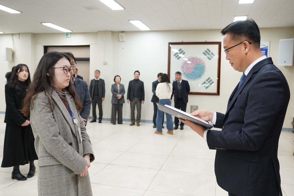 2026년 상반기 4급이하 정기인사로 정장을 입은 남성이 실내 행사장에서 한 여성에게 상장을 전달하며 내용을 읽고 있고, 여성은 긴 파마머리에 밝은 회색과 베이지가 썩힌 체크 코드를 입고 사원증을 착용한 채 두 손을 모은 상태로 서 있으며 뒤쪽에는 참석자들이 줄지어 서서 행사를 지켜보고 있다.