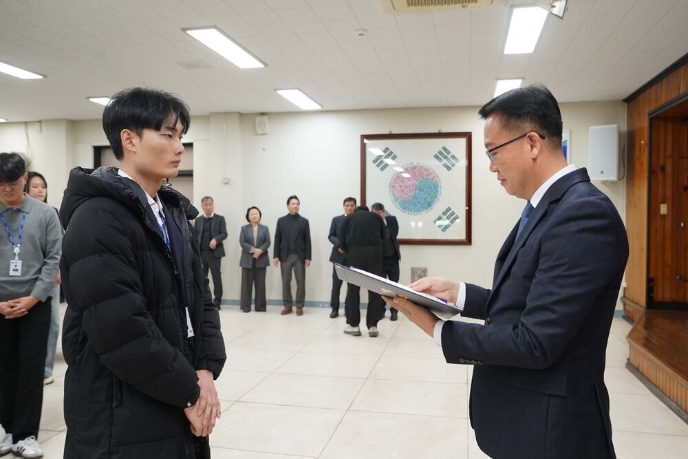 2026년 상반기 4급이하 정기인사로 정장을 입은 남성이 실내 행사장에서 한 남성에게 상장을 전달하며 내용을 읽고 있고, 남성은 검은 패딩 점퍼를 입은 채 두 손을 모은 상태로 서 있으며 뒤쪽에는 참석자들이 줄지어 서서 행사를 지켜보고 있다.