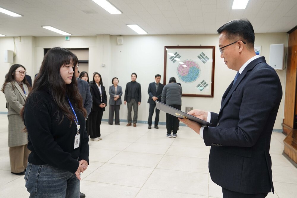 2026년 상반기 4급이하 정기인사로 정장을 입은 남성이 실내 행사장에서 한 여성에게 상장을 전달하며 내용을 읽고 있고, 여성은 검은 상의를 입고 두 손을 모은 상태로 서 있으며 뒤쪽에는 참석자들이 줄지어 서서 행사를 지켜보고 있다.