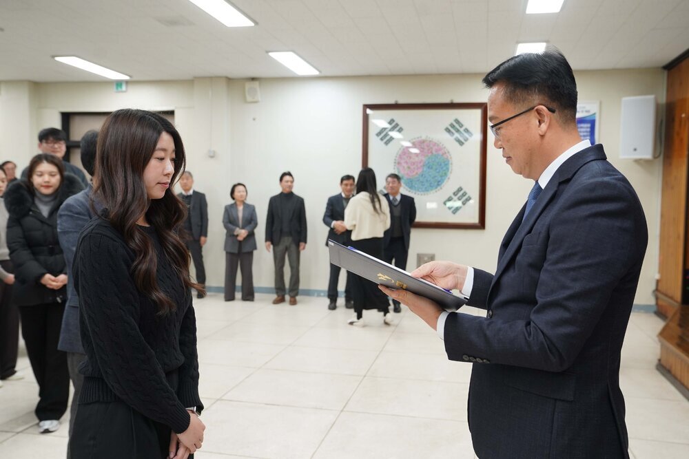2026년 상반기 4급이하 정기인사로 정장을 입은 남성이 실내 행사장에서 한 여성에게 상장을 전달하며 내용을 읽고 있고, 여성은 검은 상의를 입고 두 손을 모은 상태로 서 있으며 뒤쪽에는 참석자들이 줄지어 서서 행사를 지켜보고 있다.