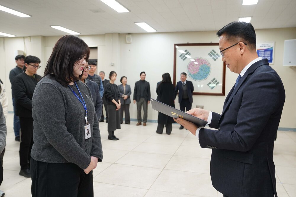 2026년 상반기 4급이하 정기인사로 정장을 입은 남성이 실내 행사장에서 한 여성에게 상장을 전달하며 내용을 읽고 있고, 여성은 회색 니트를 입고 사원증을 착용한 채 두 손을 모은 상태로 서 있으며 뒤쪽에는 참석자들이 줄지어 서서 행사를 지켜보고 있다.