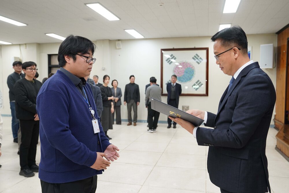 2026년 상반기 4급이하 정기인사로 정장을 입은 남성이 실내 행사장에서 한 남성에게 상장을 전달하며 내용을 읽고 있고, 남성은 파란 니트를 입고 사원증을 착용한 채 두 손을 모은 상태로 서 있으며 뒤쪽에는 참석자들이 줄지어 서서 행사를 지켜보고 있다.