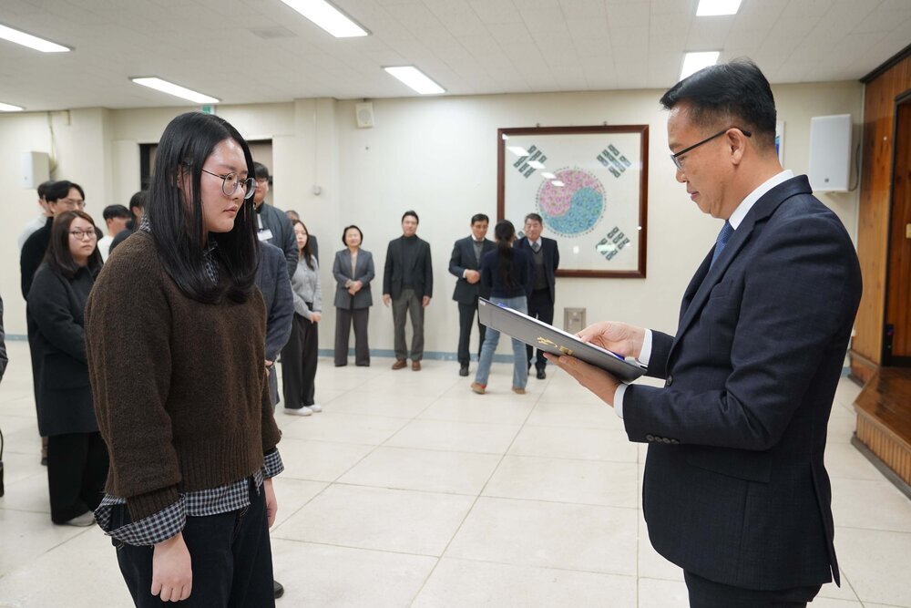2026년 상반기 4급이하 정기인사로 정장을 입고 안경을 쓴 남성이 실내 행사장에서 한 여성에게 상장을 전달하며 내용을 읽고 있고, 여성은 갈색 니트와 안경을 착용한 채 두 손을 모은 채 서 있으며 뒤쪽에는 참석자들이 줄지어 서서 행사를 지켜보고 있다.
