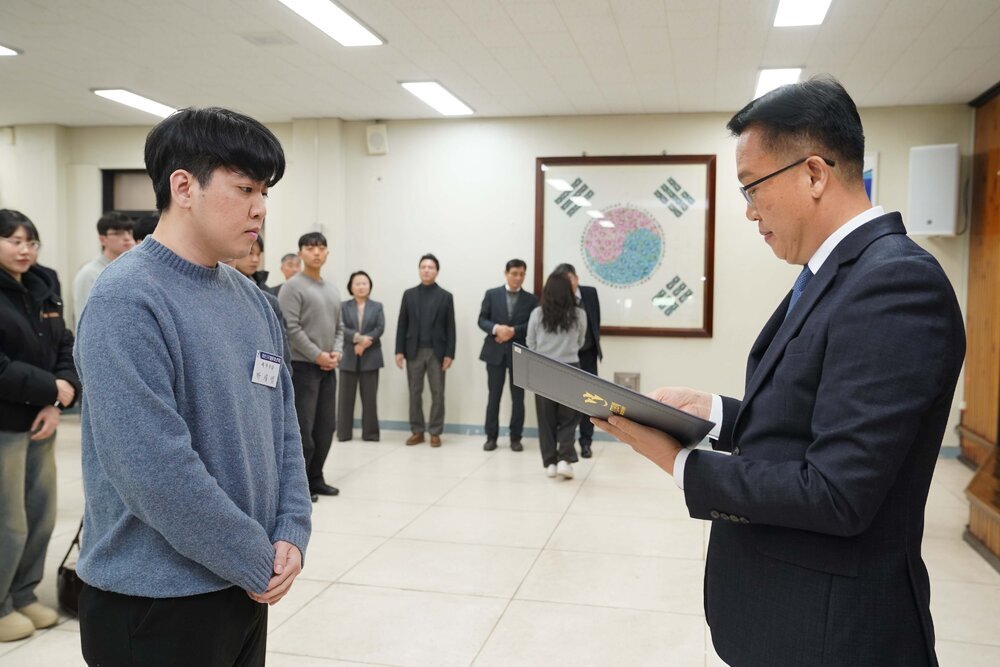 2026년 상반기 4급이하 정기인사로 정장을 입고 안경을 쓴 남성이 실내 행사장에서 한 남성에게 상장을 전달하며 내용을 읽고 있고, 남성은 회색 니트를 입고 두 손을 모은 채 서 있으며 뒤쪽에는 참석자들이 줄지어 서서 행사를 지켜보고 있다.