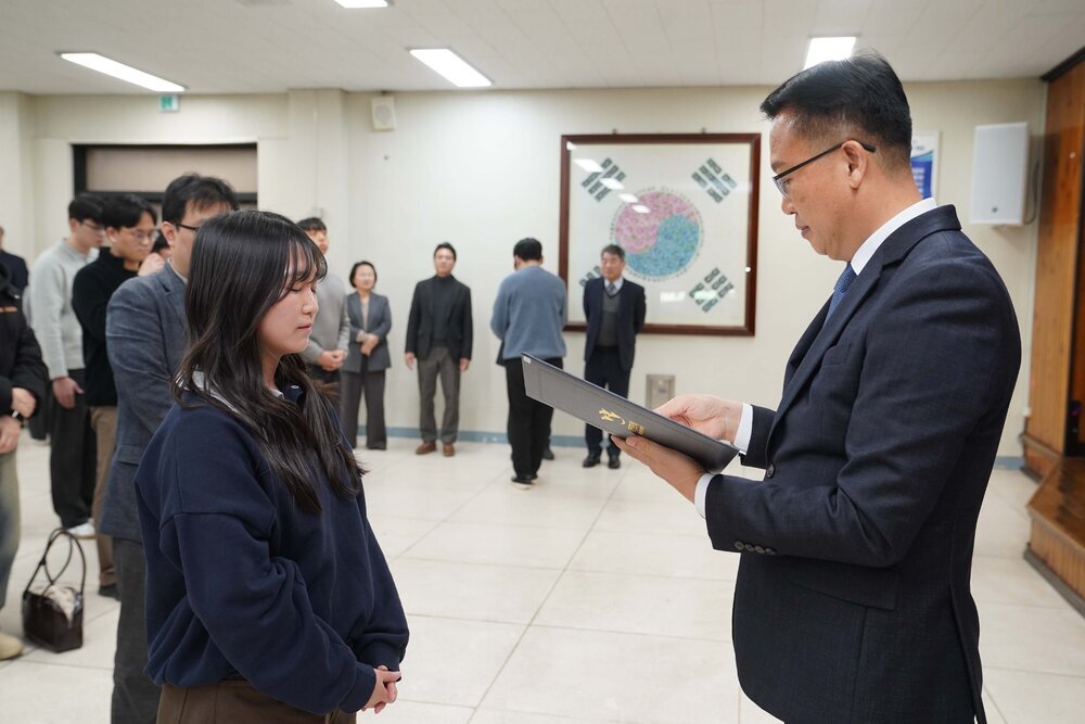 2026년 상반기 4급이하 정기인사로 정장을 입고 안경을 쓴 남성이 실내 행사장에서 한 여성에게 상장을 전달하며 내용을 읽고 있고, 여성은 남색 맨투맨을 입고 두 손을 모은 채 서 있으며 뒤쪽에는 참석자들이 줄지어 서서 행사를 지켜보고 있다.