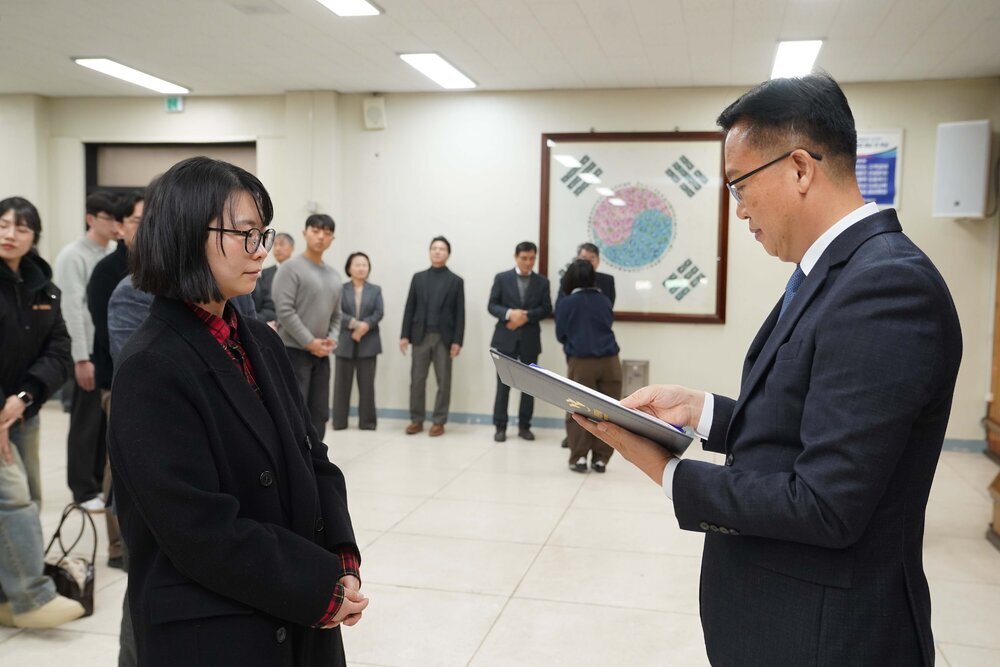 2026년 상반기 4급이하 정기인사로 정장을 입고 안경을 쓴 남성이 실내 행사장에서 한 여성에게 상장을 전달하며 내용을 읽고 있고, 여성은 검은 코트와 안경을 착용한 채 두 손을 모은 채 서 있으며 뒤쪽에는 참석자들이 줄지어 서서 행사를 지켜보고 있다.