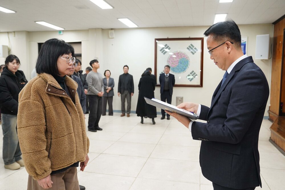 2026년 상반기 4급이하 정기인사로 정장을 입고 안경을 쓴 남성이 실내 행사장에서 한 여성에게 상장을 전달하며 내용을 읽고 있고, 여성은 갈색 퍼 재킷을 입고 안경을 착용한 채 두 손을 모은 채 서 있으며 뒤쪽에는 참석자들이 줄지어 서서 행사를 지켜보고 있다.