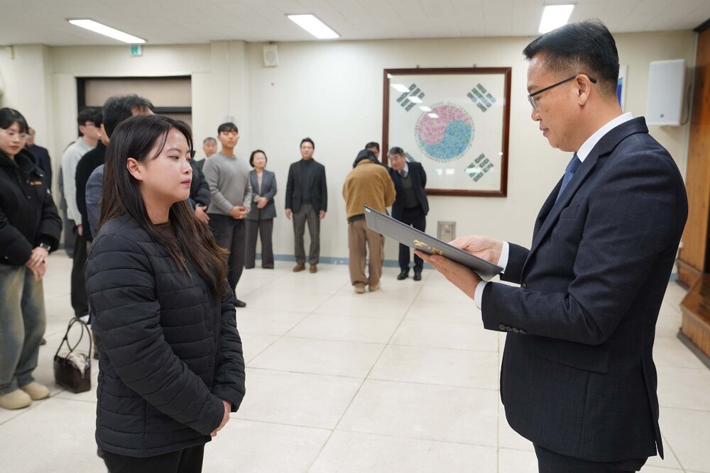 2026년 상반기 4급이하 정기인사로 정장을 입고 안경을 쓴 남성이 실내 행사장에서 한 여성에게 상장을 전달하며 내용을 읽고 있고, 여성은 검은 패딩을 입고 두 손을 모은 채 서 있으며 뒤쪽에는 참석자들이 줄지어 서서 행사를 지켜보고 있다.