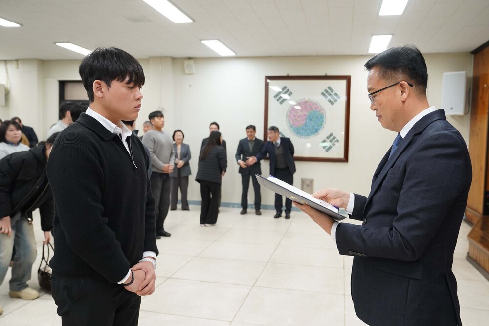 2026년 상반기 4급이하 정기인사로 정장을 입고 안경을 쓴 남성이 실내 행사장에서 한 남성에게 상장을 전달하며 내용을 읽고 있고, 남성은 검은 니트를 입고 두 손을 모은 채 서 있으며 뒤쪽에는 참석자들이 줄지어 서서 행사를 지켜보고 있다.
