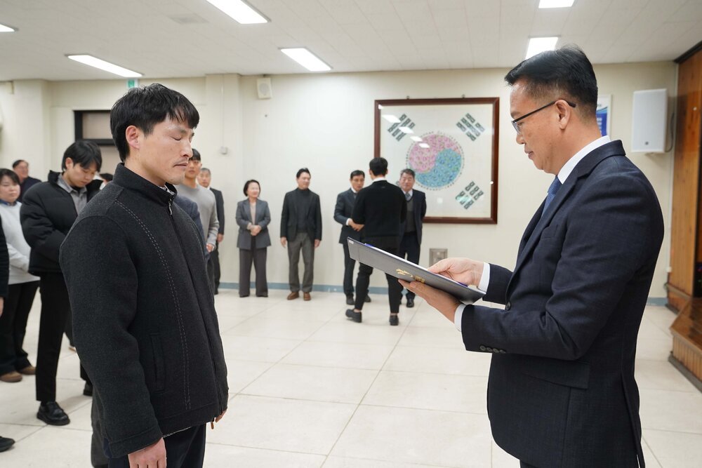 2026년 상반기 4급이하 정기인사로 정장을 입고 안경을 쓴 남성이 실내 행사장에서 한 남성에게 상장을 전달하며 내용을 읽고 있고, 남성은 검은 집업 상의를 입고 서 있으며 뒤쪽에는 참석자들이 줄지어 서서 행사를 지켜보고 있다.
