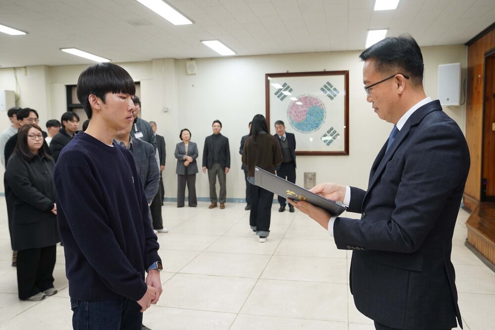 2026년 상반기 4급이하 정기인사로 정장을 입고 안경을 쓴 남성이 실내 행사장에서 한 남성에게 상장을 전달하며 내용을 읽고 있고, 남성은 짙은 색 니트를 입고 두 손을 모은 채 서 있으며 뒤쪽에는 참석자들이 줄지어 서서 행사를 지켜보고 있다.