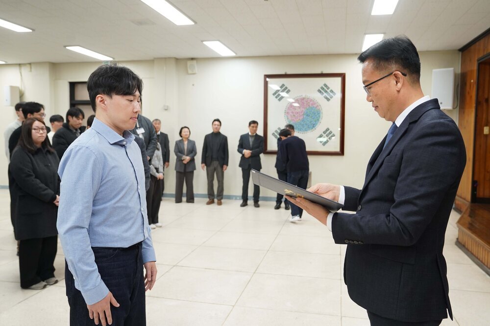 2026년 상반기 4급이하 정기인사로 정장을 입고 안경을 쓴 남성이 실내 행사장에서 한 남성에게 상장을 전달하며 내용을 읽고 있고, 남성은 파란 셔츠를 입고 서 있으며 뒤쪽에는 참석자들이 줄지어 서서 행사를 지켜보고 있다.