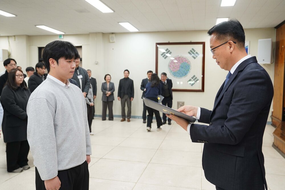 2026년 상반기 4급이하 정기인사로 정장을 입고 안경을 쓴 남성이 실내 행사장에서 한 남성에게 상장을 전달하며 내용을 읽고 있고, 남성은 회색 맨투맨을 입고 서 있으며 뒤쪽에는 참석자들이 줄지어 서서 행사를 지켜보고 있다.