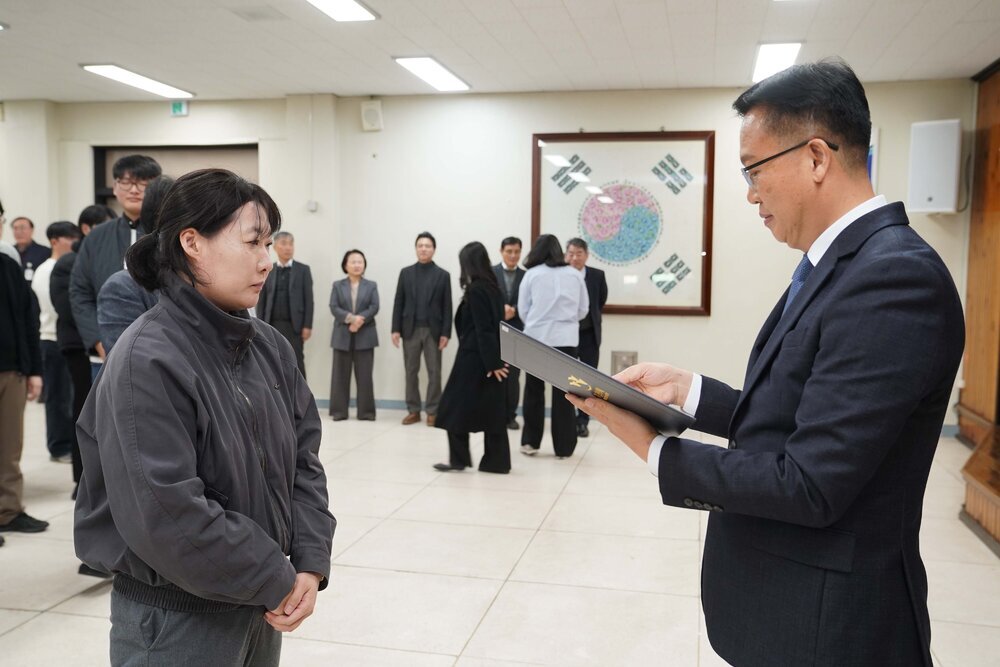 2026년 상반기 4급이하 정기인사로 정장을 입고 안경을 쓴 남성이 실내 행사장에서 한 여성에게 상장을 전달하며 내용을 읽고 있고, 여성은 회색 점퍼를 입고 두 손을 모은 채 서 있으며 뒤쪽에는 참석자들이 줄지어 서서 행사를 지켜보고 있다.
