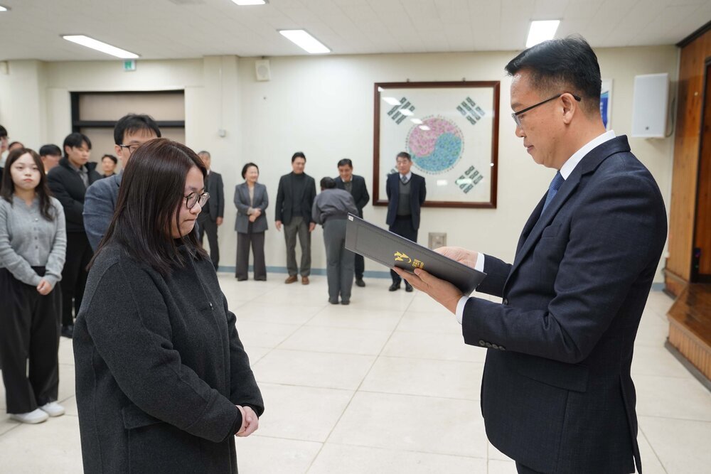 2026년 상반기 4급이하 정기인사로 정장을 입고 안경을 쓴 남성이 실내 행사장에서 한 여성에게 상장을 전달하며 내용을 읽고 있고, 여성은 검은 코트를 입고 안경을 착용한 채 두 손을 모은 채 서 있으며 뒤쪽에는 참석자들이 줄지어 서서 행사를 지켜보고 있다.