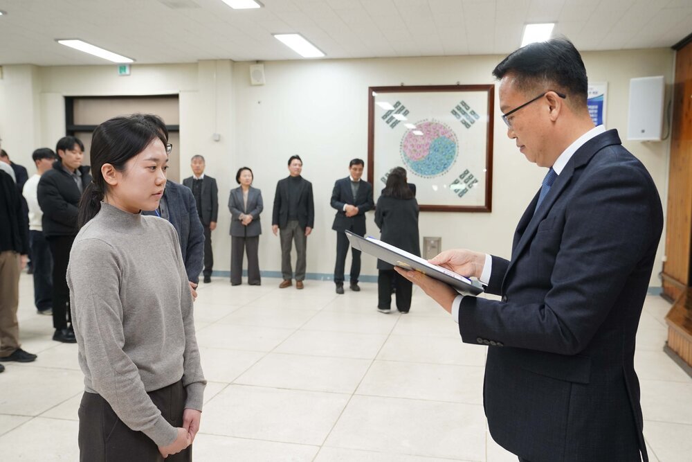 2026년 상반기 4급이하 정기인사로 정장을 입고 안경을 쓴 남성이 실내 행사장에서 한 여성에게 상장을 전달하며 내용을 읽고 있고, 여성은 회색 니트를 입고 두 손을 모은 채 서 있으며 뒤쪽에는 참석자들이 줄지어 서서 행사를 지켜보고 있다.