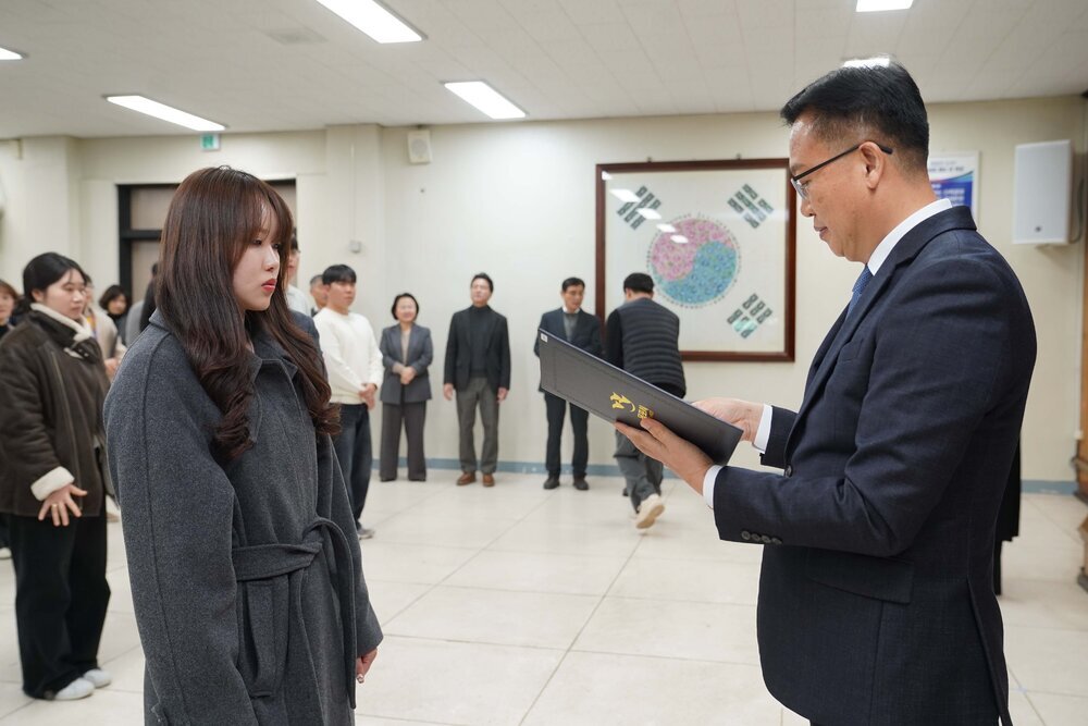 2026년 상반기 4급이하 정기인사로 정장을 입고 안경을 쓴 남성이 실내 행사장에서 한 여성에게 상장을 전달하며 내용을 읽고 있고, 여성은 회색 코트를 입고 서 있으며 뒤쪽에는 참석자들이 줄지어 서서 행사를 지켜보고 있다.