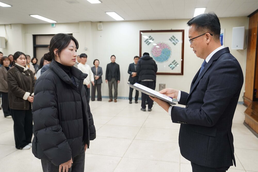 2026년 상반기 4급이하 정기인사로 정장을 입고 안경을 쓴 남성이 실내 행사장에서 한 여성에게 상장을 전달하며 내용을 읽고 있고, 여성은 검은 패딩을 입고 두 손을 모은 채 서 있으며 뒤쪽에는 참석자들이 줄지어 서서 행사를 지켜보고 있다.