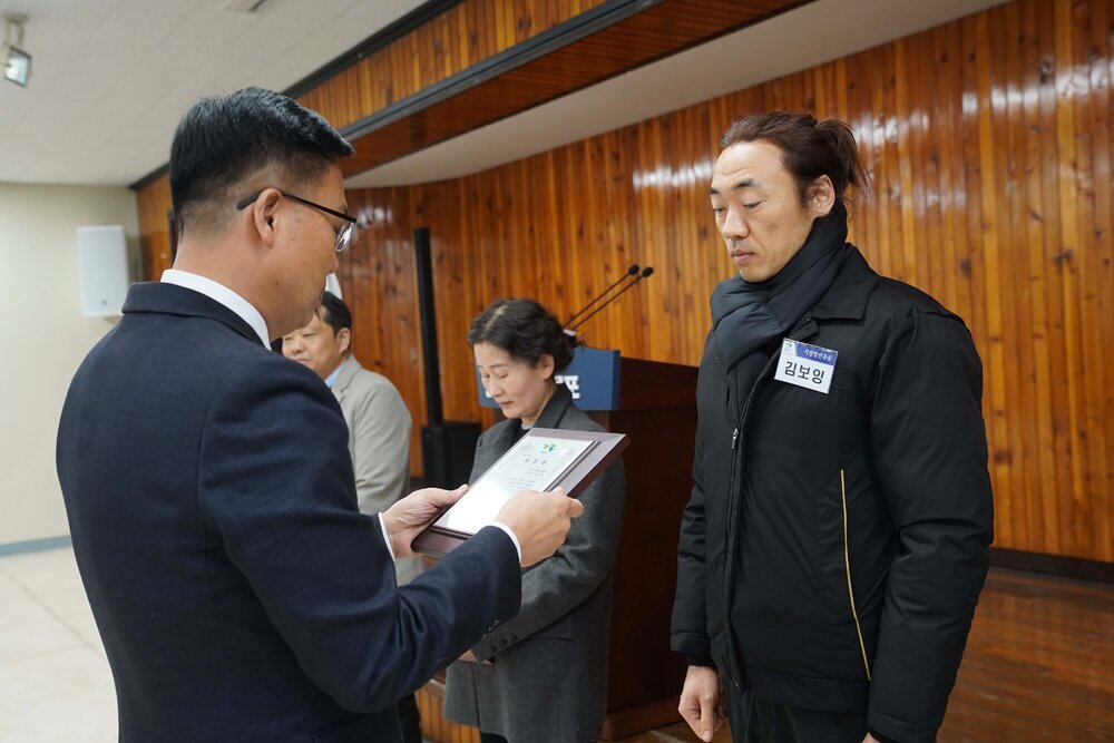 2월 정례조회에 정장을 입고 안경을 낀 남성이 머리를 뒤로 묶은 검은색아웃터에 검은색 목도리를 한 김보잉에게 패를 수여하고 있다.