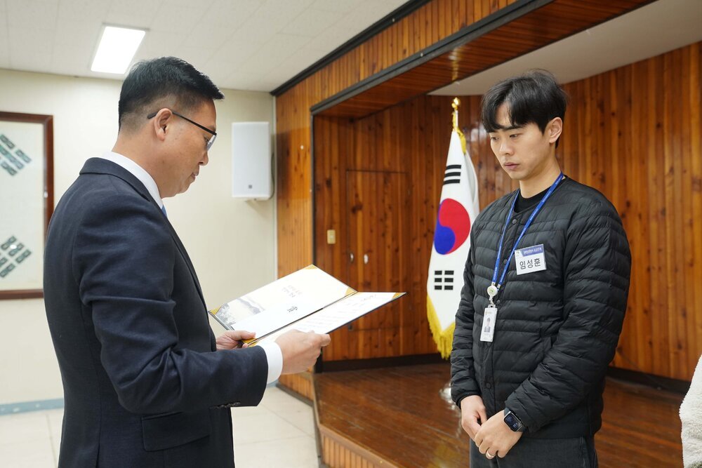 2월 정례조회에 정장을 입고 안경을 낀 남성이 검은색바지에 검은색 경랼패딩을 입은 임성훈에게 패를 수여하고 있다.