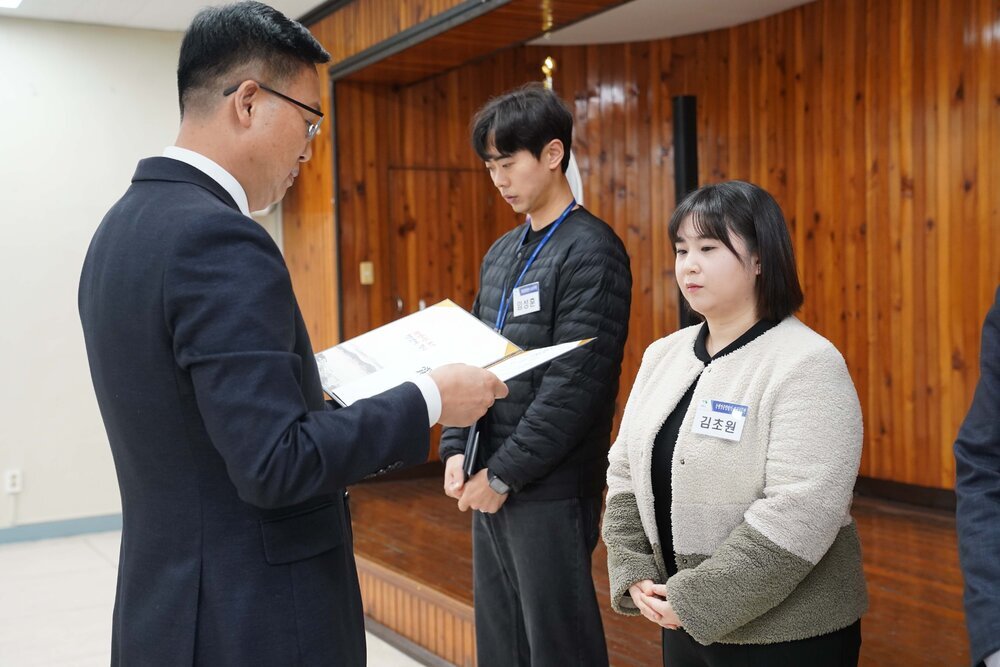 2월 정례조회에 정장을 입고 안경을 낀 남성이 베이지색, 카키색 짧은 양털자켓을 입은 김초원에게 패를 수여하고 있다.