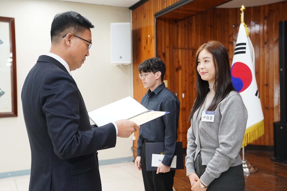 2월 정례조회에 정장을 입고 안경을 낀 남성이 베이지 목티에 회색가디건을 입은 은미경에게 패를 수여하고 있다.