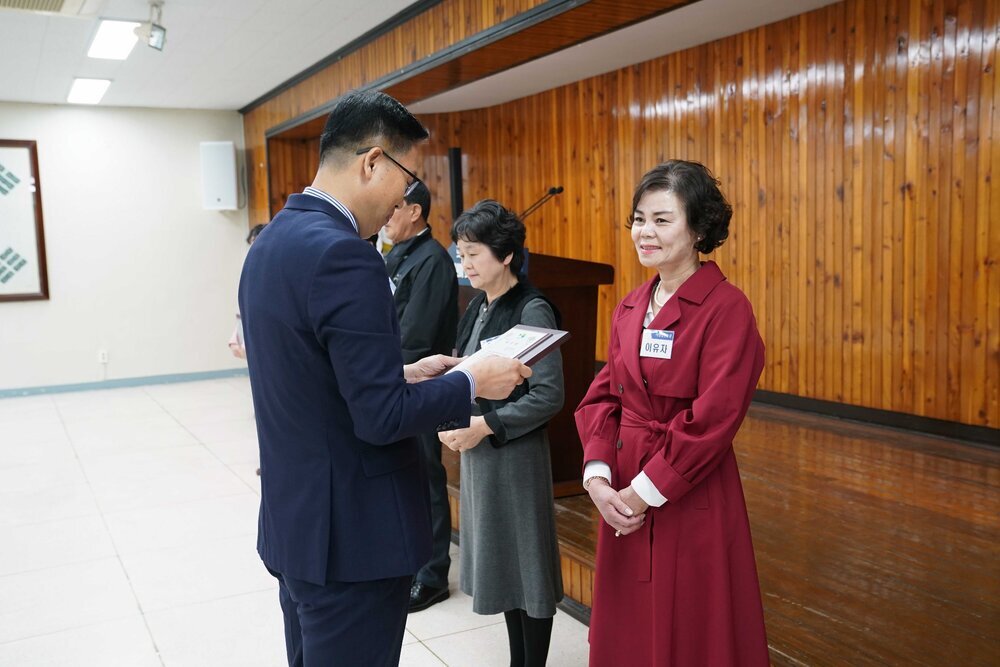 4월 정례조회에 정장을 입고 안경을 낀 남성이 와인색 원피스를 입은 이유자에게 패를 수여하고 있다
