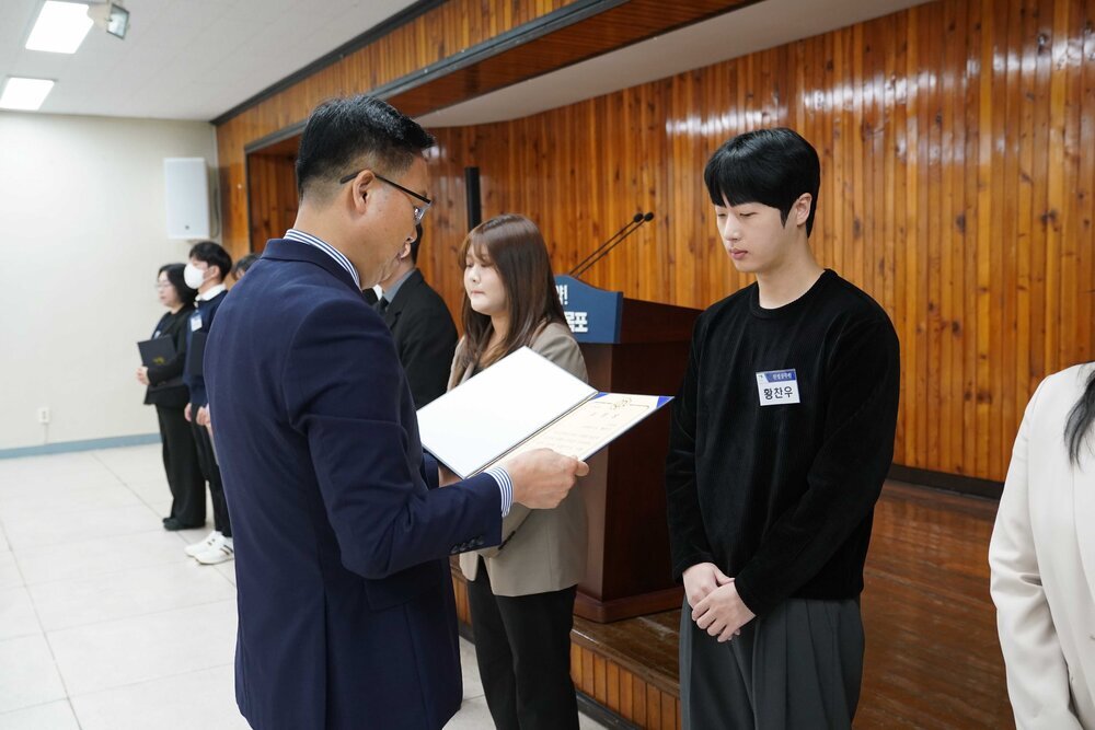 4월 정례조회에 정장을 입고 안경을 낀 남성이 검정색 스웨터를 입은 황찬우에게 패를 수여하고 있다