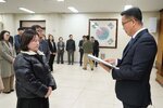 2026년 상반기 4급이하 정기인사로 정장을 입은 남성이 실내 행사장에서 한 여성에게 상장을 전달하며 내용을 읽고 있고, 여성은 검은색 짧은 패딩을 입고 두 손을 모은 상태로 서 있으며 뒤쪽으로 참석자들이 줄지어 서서 행사를 지켜보고 있다.