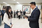 2026년 상반기 4급이하 정기인사로 정장을 입은 남성이 실내 행사장에서 한 여성에게 상장을 전달하며 내용을 읽고 있고, 여성은 긴 파마머리에 청바지와 흰색 꽈베기 니트를 입고 사원증을 착용한 채 두 손을 모은 상태로 서 있으며 뒤쪽으로 참석자들이 줄지어 서서 행사를 지켜보고 있다.