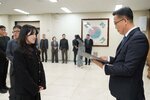 2026년 상반기 4급이하 정기인사로 정장을 입은 남성이 실내 행사장에서 한 여성에게 상장을 전달하며 내용을 읽고 있고, 여성은 긴 파마머리에 검은색 코트를 입고 두 손을 모은 상태로 서 있으며 뒤쪽에는 참석자들이 줄지어 서서 행사를 지켜보고 있다.