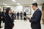 2026년 상반기 4급이하 정기인사로 정장을 입은 남성이 실내 행사장에서 한 여성에게 상장을 전달하며 내용을 읽고 있고, 여성은 어꺠 까지 오는 단발머리에 흰셔츠 검은색 자켓을 입고 사원증을 착용한 채 두 손을 모은 상태로 서 있으며 뒤쪽에는 참석자들이 줄지어 서서 행사를 지켜보고 있다.
