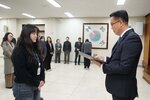2026년 상반기 4급이하 정기인사로 정장을 입은 남성이 실내 행사장에서 한 여성에게 상장을 전달하며 내용을 읽고 있고, 여성은 검은 상의를 입고 두 손을 모은 상태로 서 있으며 뒤쪽에는 참석자들이 줄지어 서서 행사를 지켜보고 있다.