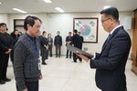 2026년 상반기 4급이하 정기인사로 정장을 입은 남성이 실내 행사장에서 한 남성에게 상장을 전달하며 내용을 읽고 있고, 남성은 회색 니트와 사원증을 착용한 채 두 손을 모은 상태로 서 있으며 뒤쪽에는 참석자들이 줄지어 서서 행사를 지켜보고 있다.