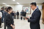 2026년 상반기 4급이하 정기인사로 정장을 입은 남성이 실내 행사장에서 한 여성에게 상장을 전달하며 내용을 읽고 있고, 여성은 검은 니트를 입고 두 손을 모은 상태로 서 있으며 뒤쪽에는 참석자들이 줄지어 서서 행사를 지켜보고 있다.