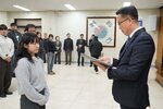 2026년 상반기 4급이하 정기인사로 정장을 입은 남성이 실내 행사장에서 한 여성에게 상장을 전달하며 내용을 읽고 있고, 여성은 회색 니트를 입고 두 손을 모은 상태로 서 있으며 뒤쪽에는 참석자들이 줄지어 서서 행사를 지켜보고 있다.