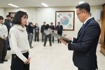 2026년 상반기 4급이하 정기인사로 정장을 입은 남성이 실내 행사장에서 한 여성에게 상장을 전달하며 내용을 읽고 있고, 여성은 아이보리색 니트를 입고 두 손을 모은 상태로 서 있으며 뒤쪽에는 참석자들이 줄지어 서서 행사를 지켜보고 있다.