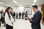 2026년 상반기 4급이하 정기인사로 정장을 입은 남성이 실내 행사장에서 한 여성에게 상장을 전달하며 내용을 읽고 있고, 여성은 밝은 색 후드티를 입고 두 손을 모은 상태로 서 있으며 뒤쪽에는 참석자들이 줄지어 서서 행사를 지켜보고 있다.