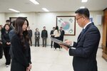 2026년 상반기 4급이하 정기인사로 정장을 입은 남성이 실내 행사장에서 한 여성에게 상장을 전달하며 내용을 읽고 있고, 여성은 검은 상의를 입고 두 손을 모은 상태로 서 있으며 뒤쪽에는 참석자들이 줄지어 서서 행사를 지켜보고 있다.