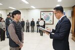 2026년 상반기 4급이하 정기인사로 정장을 입은 남성이 실내 행사장에서 한 남성에게 상장을 전달하며 내용을 읽고 있고, 남성은 검은 패딩 조끼를 입은 채 두 손을 모은 상태로 서 있으며 뒤쪽에는 참석자들이 줄지어 서서 행사를 지켜보고 있다.