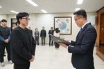 2026년 상반기 4급이하 정기인사로 정장을 입은 남성이 실내 행사장에서 한 남성에게 상장을 전달하며 내용을 읽고 있고, 남성은 검은 외투를 입고 안경을 착용한 채 두 손을 모은 상태로 서 있으며 뒤쪽에는 참석자들이 줄지어 서서 행사를 지켜보고 있다.