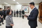 2026년 상반기 4급이하 정기인사로 정장을 입은 남성이 실내 행사장에서 한 여성에게 상장을 전달하며 내용을 읽고 있고, 여성은 회색 니트를 입고 두 손을 모은 상태로 서 있으며 뒤쪽에는 참석자들이 줄지어 서서 행사를 지켜보고 있다.