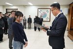 2026년 상반기 4급이하 정기인사로 정장을 입고 안경을 쓴 남성이 실내 행사장에서 한 여성에게 상장을 전달하며 내용을 읽고 있고, 여성은 남색 니트를 입고 두 손을 모은 채 서 있으며 뒤쪽에는 참석자들이 줄지어 서서 행사를 지켜보고 있다.