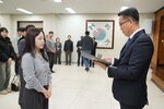 2026년 상반기 4급이하 정기인사로 정장을 입고 안경을 쓴 남성이 실내 행사장에서 한 여성에게 상장을 전달하며 내용을 읽고 있고, 여성은 회색 니트를 입고 두 손을 모은 채 서 있으며 뒤쪽에는 참석자들이 줄지어 서서 행사를 지켜보고 있다.