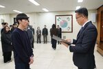 2026년 상반기 4급이하 정기인사로 정장을 입고 안경을 쓴 남성이 실내 행사장에서 한 남성에게 상장을 전달하며 내용을 읽고 있고, 남성은 짙은 색 니트를 입고 두 손을 모은 채 서 있으며 뒤쪽에는 참석자들이 줄지어 서서 행사를 지켜보고 있다.