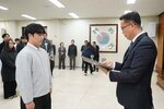 2026년 상반기 4급이하 정기인사로 정장을 입고 안경을 쓴 남성이 실내 행사장에서 한 남성에게 상장을 전달하며 내용을 읽고 있고, 남성은 회색 맨투맨을 입고 서 있으며 뒤쪽에는 참석자들이 줄지어 서서 행사를 지켜보고 있다.