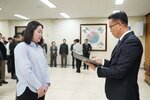 2026년 상반기 4급이하 정기인사로 정장을 입고 안경을 쓴 남성이 실내 행사장에서 한 여성에게 상장을 전달하며 내용을 읽고 있고, 여성은 밝은 색 셔츠를 입고 두 손을 모은 채 서 있으며 뒤쪽에는 참석자들이 줄지어 서서 행사를 지켜보고 있다.