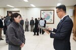 2026년 상반기 4급이하 정기인사로 정장을 입고 안경을 쓴 남성이 실내 행사장에서 한 여성에게 상장을 전달하며 내용을 읽고 있고, 여성은 회색 점퍼를 입고 두 손을 모은 채 서 있으며 뒤쪽에는 참석자들이 줄지어 서서 행사를 지켜보고 있다.